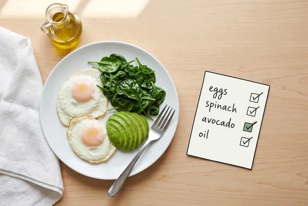 Two fried eggs with wilted spinach and sliced avocado on a white plate beside a small jug of olive oil