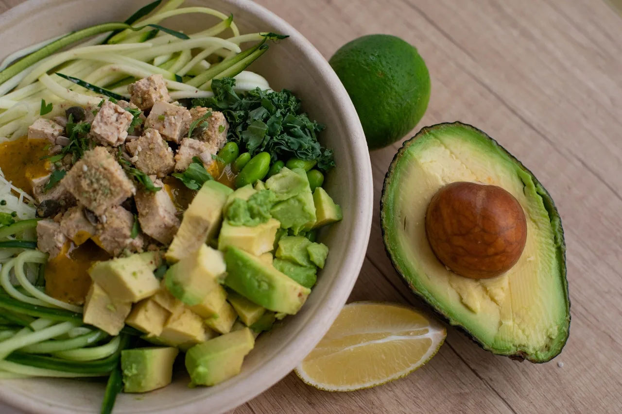 Bowl of diced chicken, avocado, zucchini noodles and herbs with lime, a whole avocado half beside it
