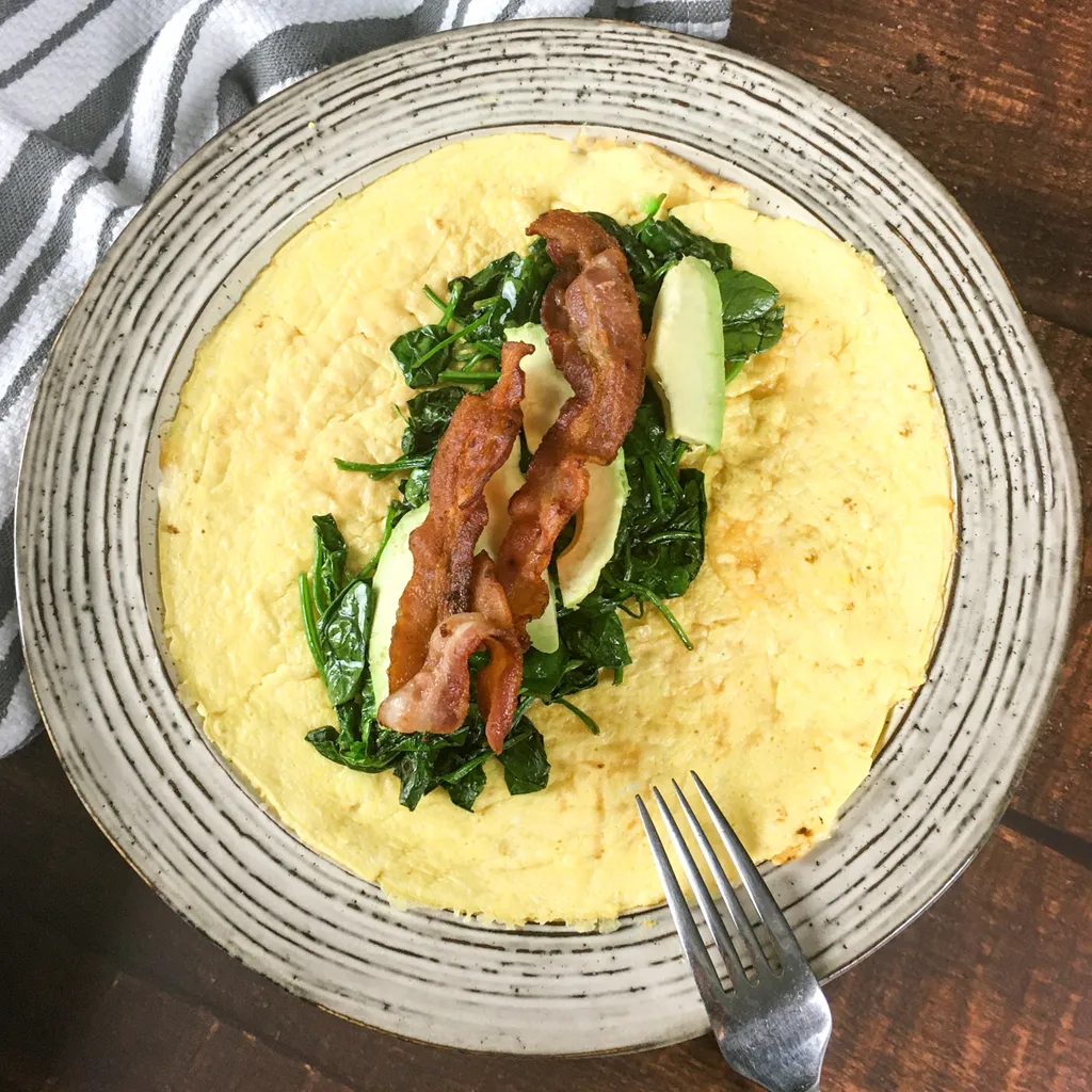 A thin golden egg wrap laid flat on a speckled stoneware plate, topped with wilted spinach, two strips of crisp bacon and slices of avocado, with a fork beside it on a dark wooden table