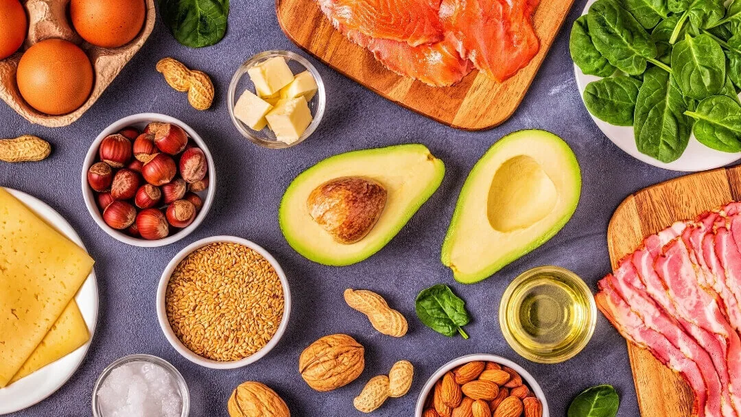 Keto-friendly foods including salmon, eggs, avocado, cheese, nuts, olive oil, and low-carb vegetables arranged on a table